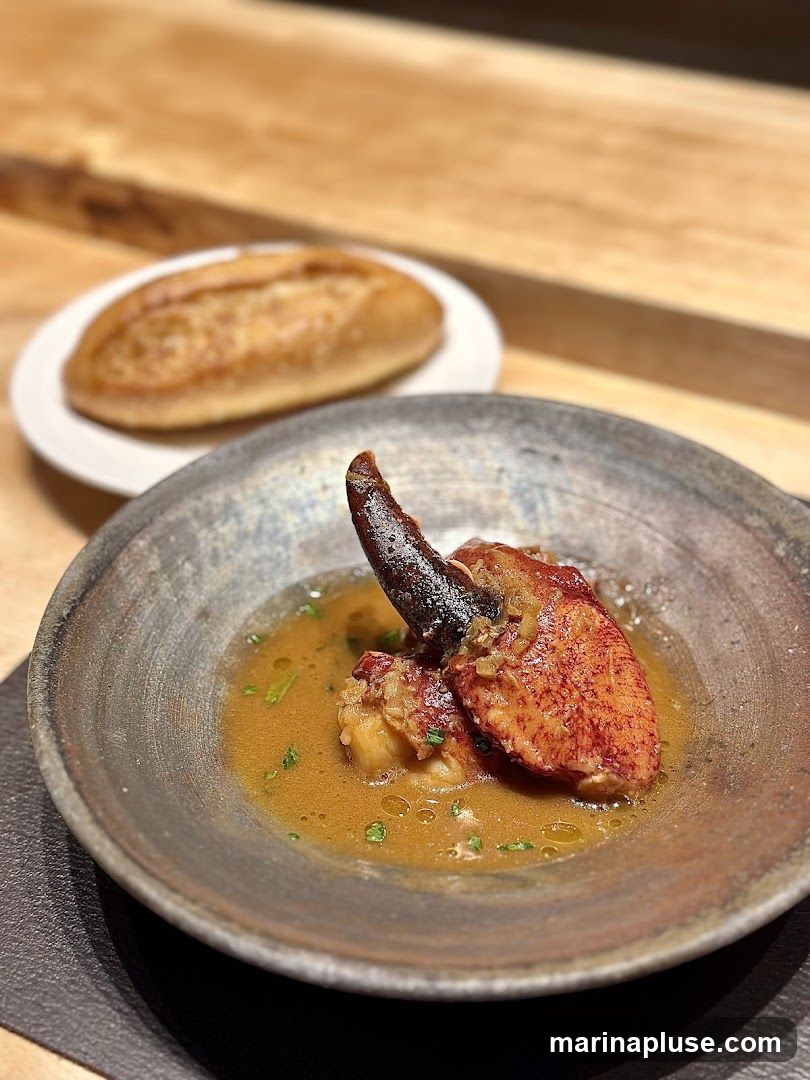 A close-up shot of a cooked lobster claw served in a rustic bowl with a light broth and garnished with chopped herbs. A small bread roll is visible on a white plate in the blurred background. The table surface appears to be made of wood.