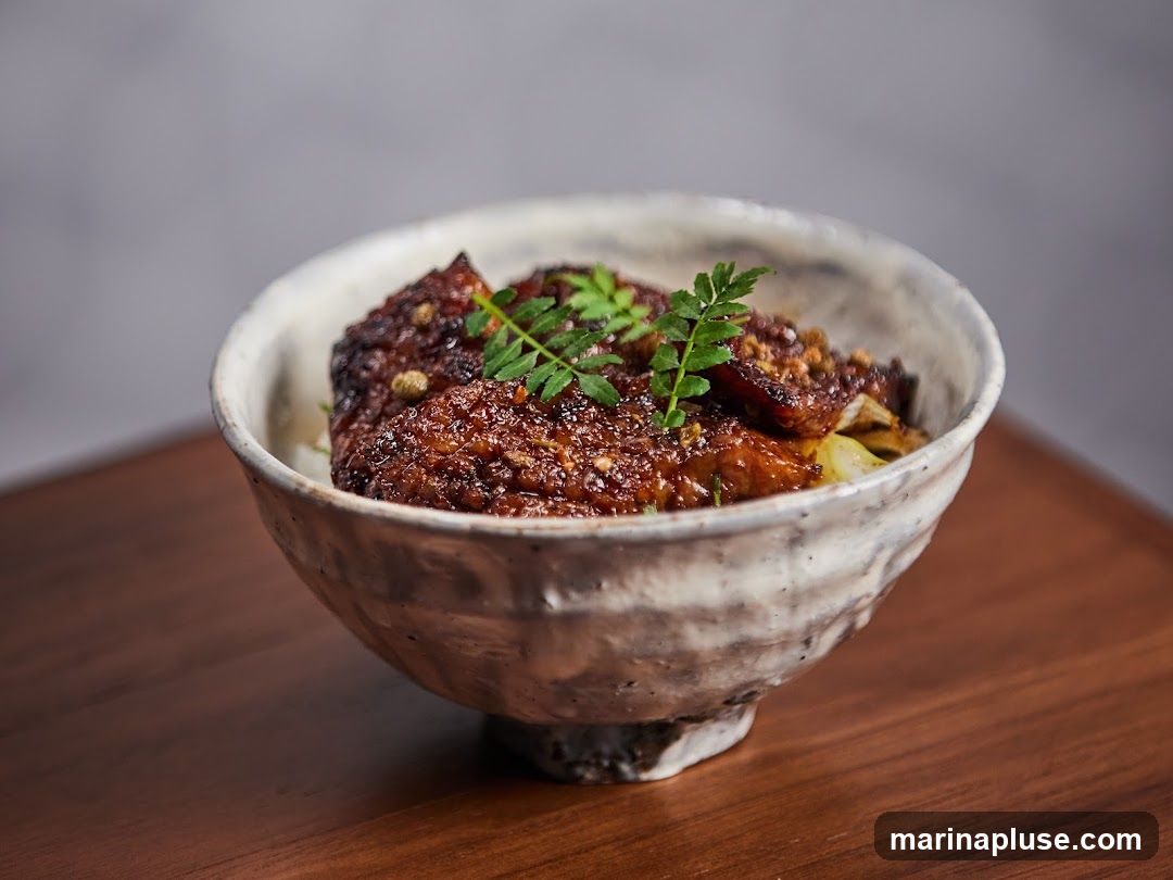 A close-up shot of a bowl of food, likely a rice bowl dish, with glazed meat and a sprig of parsley on top. The bowl is rustic and textured, and it sits on a wooden surface. The background is a plain grey.