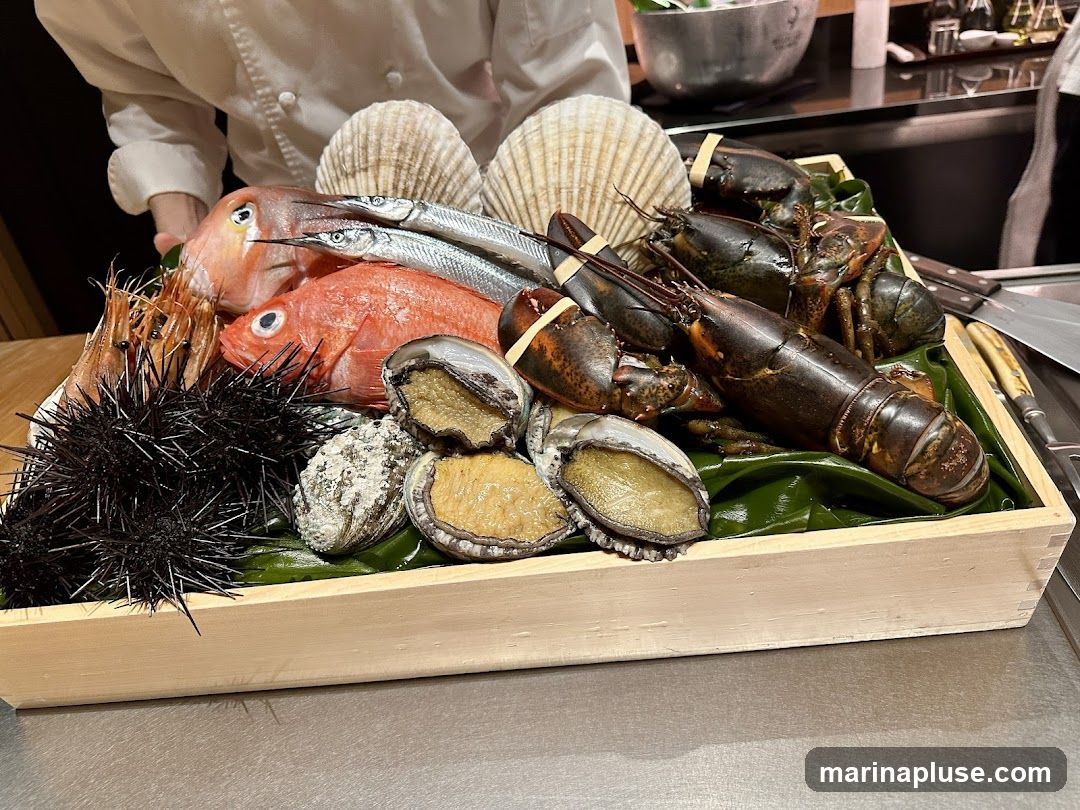 A wooden tray filled with a variety of fresh seafood, including lobsters, fish, abalone, sea urchins, shrimp, and scallops. A chef in a white uniform is visible in the background.
