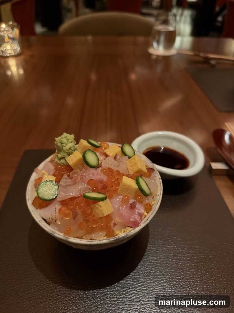 A bowl of chirashi sushi, featuring rice topped with various raw fish, ikura (salmon roe), tamagoyaki (rolled omelet), and cucumber slices. A small dish of soy sauce and a dollop of wasabi are also visible. The dish is presented on a dark placemat on a wooden table, with a blurred background of a restaurant interior.
