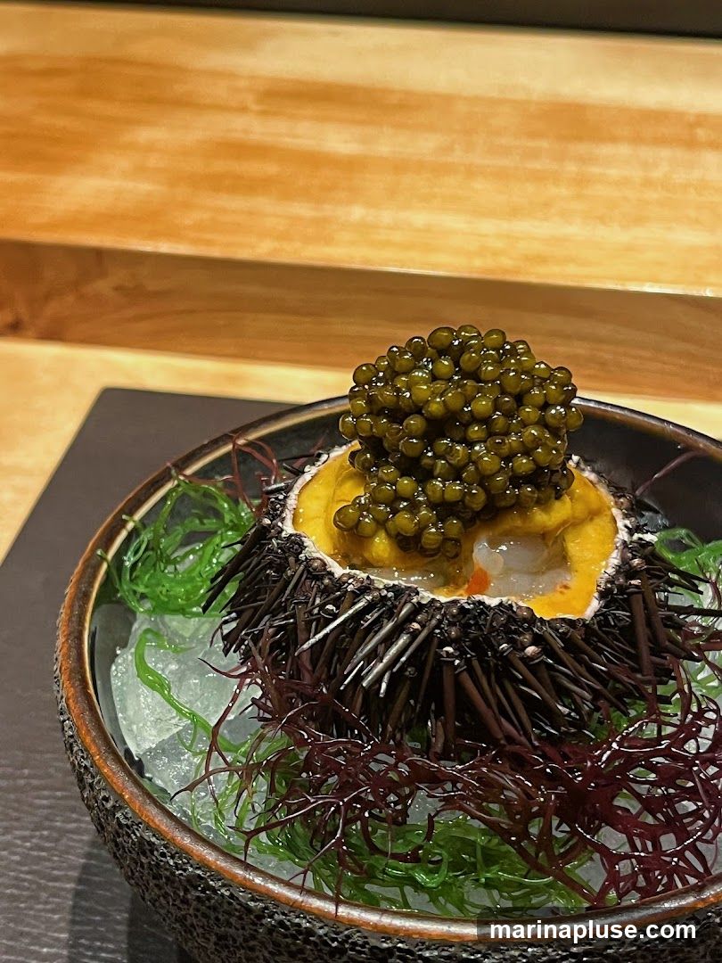 A close-up shot of a sea urchin dish. The sea urchin is filled with bright yellow uni and topped with a generous portion of dark caviar. It is served in a textured bowl filled with crushed ice and garnished with strands of red and green seaweed. The background shows a wooden surface.