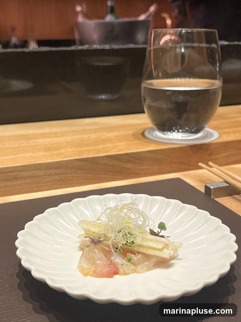 A delicate appetizer of thinly sliced raw fish, possibly sea bass or snapper, is presented on a fluted white plate. The sashimi is topped with finely shredded white vegetables, microgreens, and a light dressing. A glass of water sits to the right, with chopsticks resting on a metal chopstick holder in the background. The setting appears to be a restaurant with a wooden counter and a dark, reflective surface behind the food.