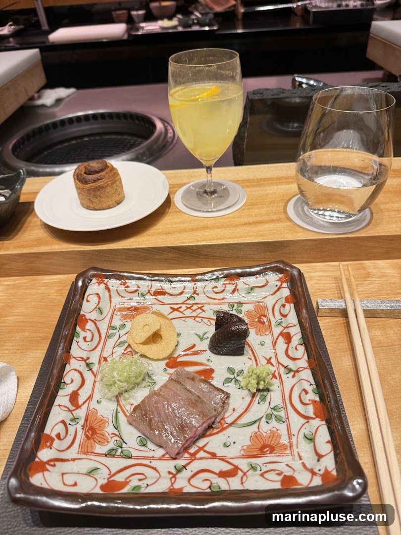 A close-up view of a small appetizer plate with a slice of meat, wasabi, and other garnishes. A glass of lemonade with a lemon slice and a glass of water are also visible, along with a small pastry on a separate plate. The setting appears to be a restaurant with a hibachi grill in the background.