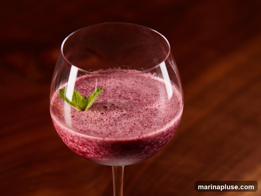 A close-up shot of a frothy, deep purple berry smoothie in a wine glass, garnished with a fresh mint leaf. The drink is served on a dark wooden surface.