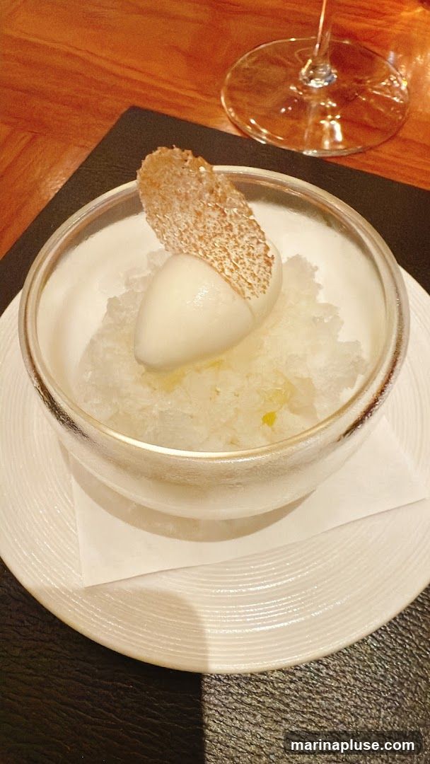 A close-up shot of a dessert served in a clear bowl. The dessert consists of shaved ice, a scoop of white ice cream, and a thin, crispy garnish. The bowl is placed on a white plate with a textured rim, which sits on a dark placemat. A wine glass stem is visible in the upper right corner, suggesting a dining setting.