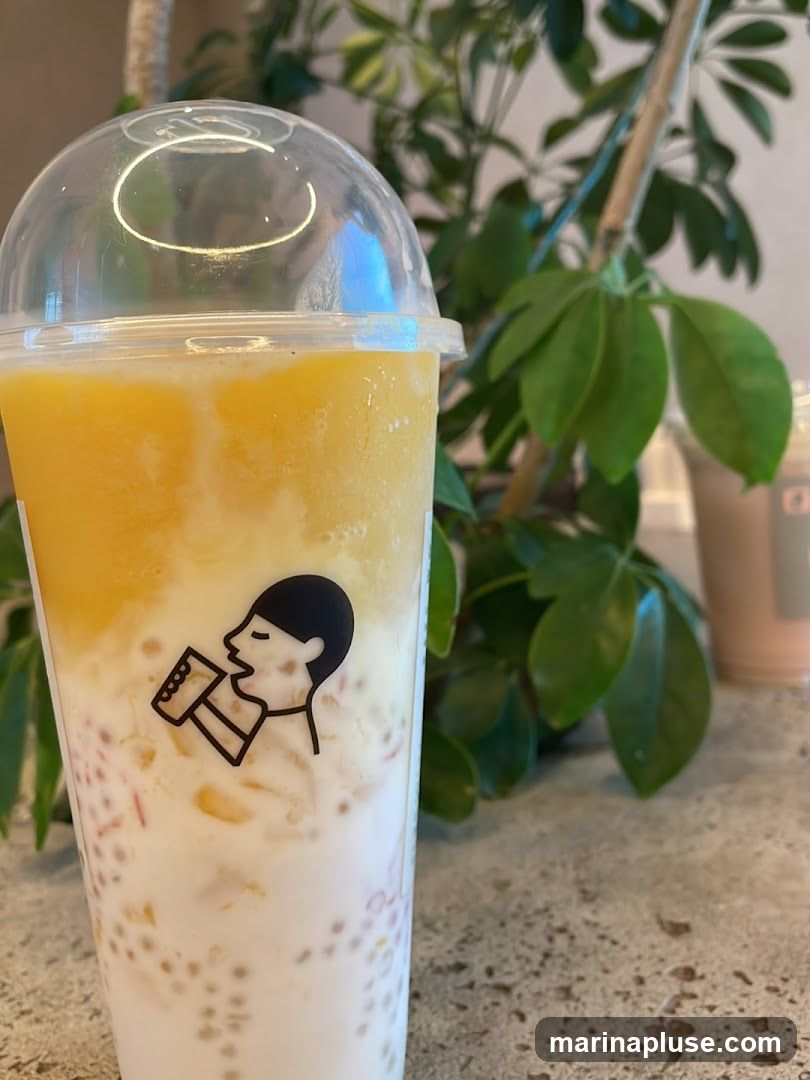 A close-up shot of a clear plastic cup filled with a layered drink. The bottom layer appears to be milk with small, round, pinkish-brown tapioca pearls and chunks of fruit. Above this is a creamy white layer, and the top layer is a thick, icy yellow mixture, likely mango. A black silhouette logo of a person drinking from a cup is printed on the side of the cup. In the background, out of focus, are green leafy plants and another drink cup.