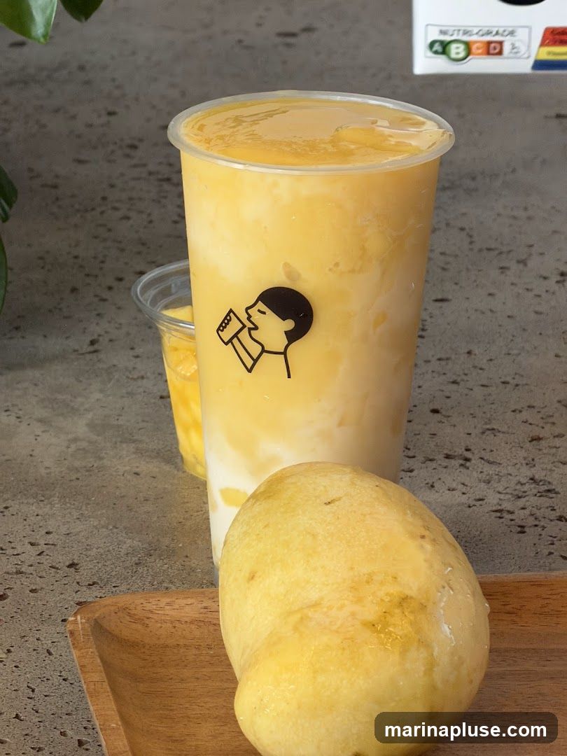 A close-up shot of a mango smoothie in a clear plastic cup, with a whole mango in the foreground on a wooden tray. The cup has a black logo of a person drinking. Another smaller cup with mango pieces is visible behind the main drink. A nutritional grade chart is visible in the upper right corner.