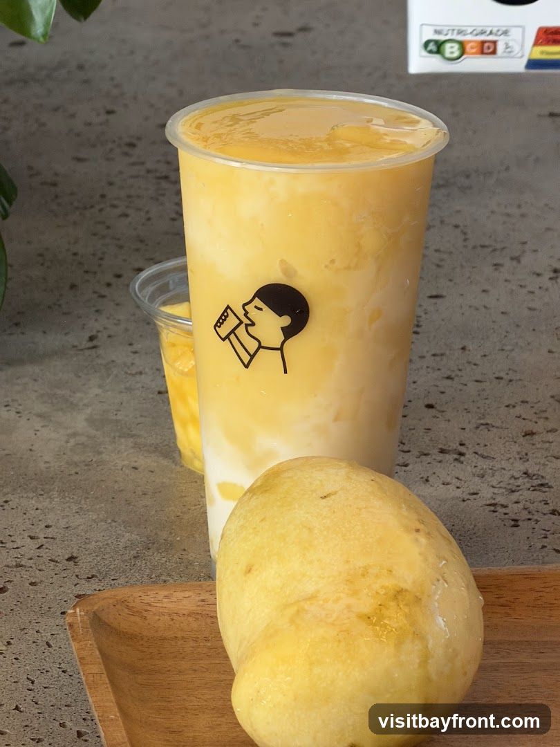 A close-up shot of a mango smoothie in a clear plastic cup, with a whole mango in the foreground on a wooden tray. The cup has a black logo of a person drinking. Another smaller cup with mango pieces is visible behind the main drink. A nutritional grade chart is visible in the upper right corner.