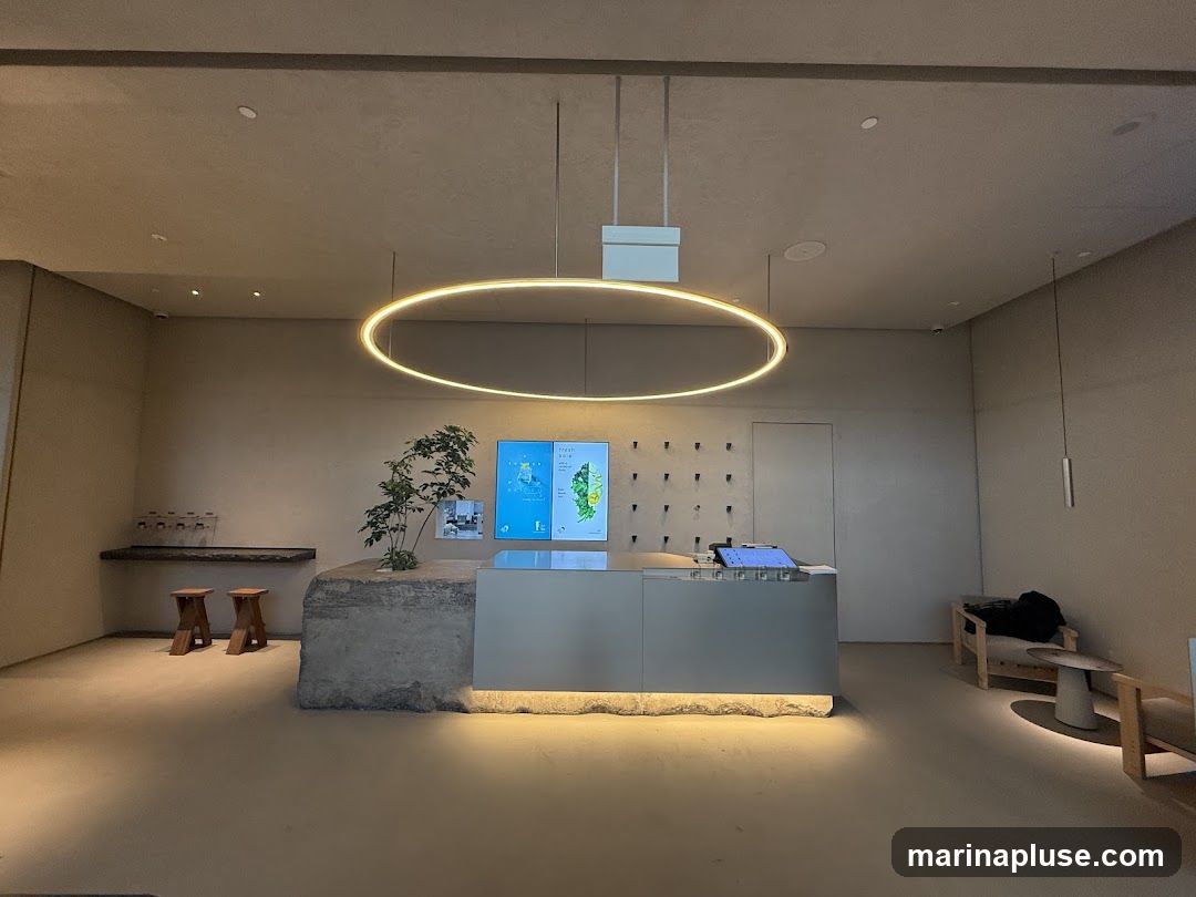 A minimalist cafe interior with a sleek counter, a large circular pendant light, and a small tree. There are stools and seating areas, and digital screens displaying information. The overall aesthetic is clean and contemporary.