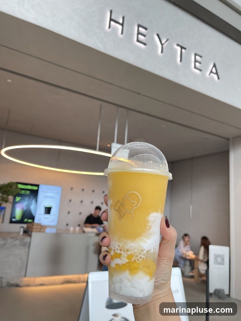A person is holding a plastic cup with a yellow and white layered drink, likely a smoothie or bubble tea, in front of a Heytea establishment. The brand's name,