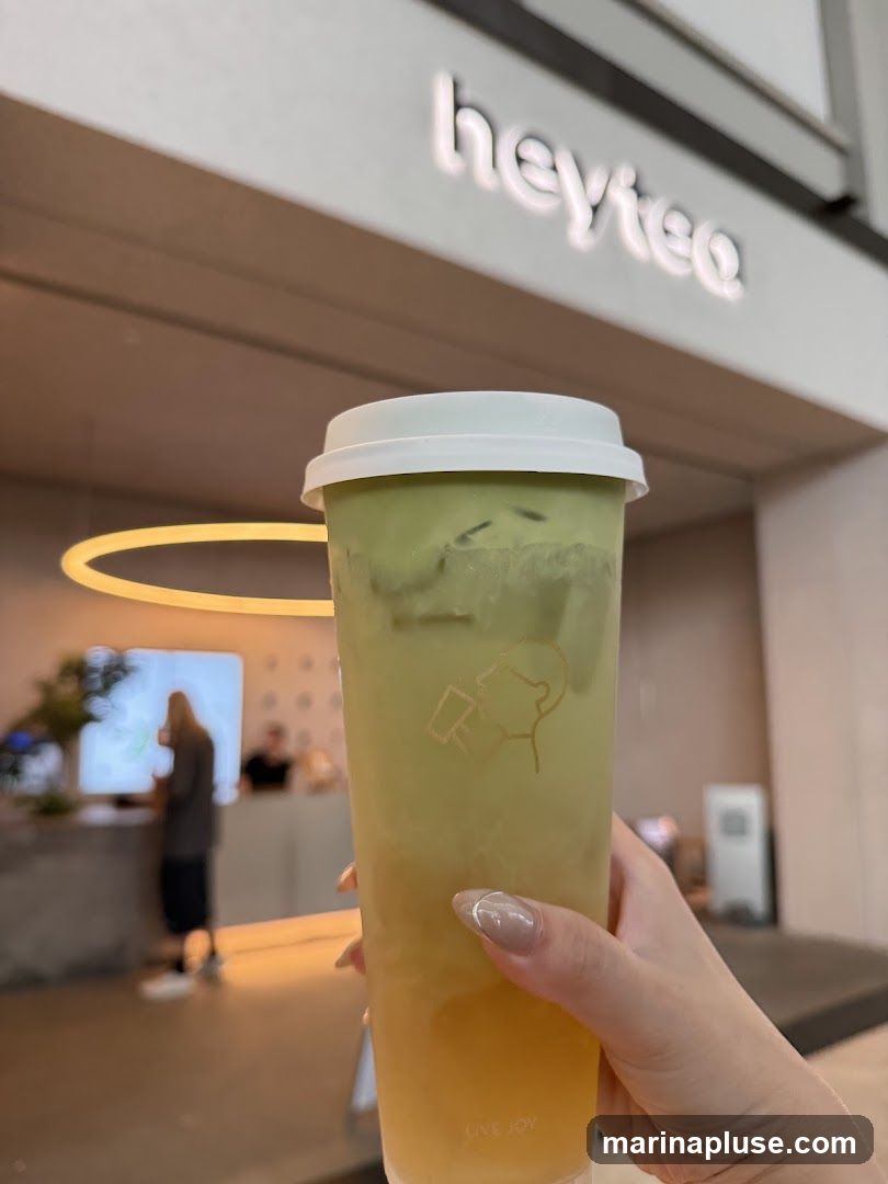 A hand holds a tall plastic cup with a white lid, filled with a layered green and yellow beverage, likely a tea drink. The cup has a faint outline logo of a person drinking. In the background, the illuminated sign "heytea" is visible above a counter area where people are blurred. The overall scene suggests an indoor setting of a beverage shop.