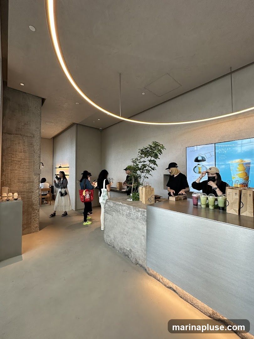 A modern cafe interior with a minimalist design. Customers are seen at the counter ordering drinks, and others are seated at tables in the background. The counter area features a sleek metallic surface and a textured concrete base. A large screen displays images of food and drinks. The lighting is soft, with a prominent curved LED strip on the ceiling.