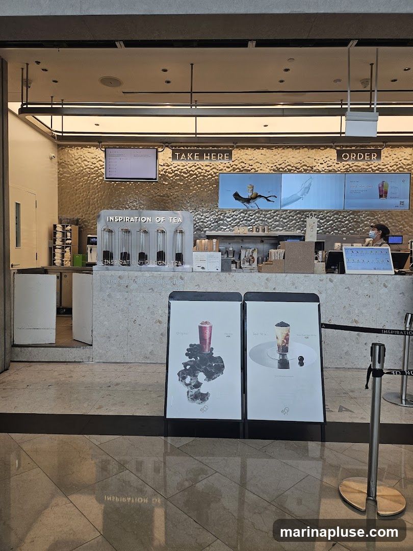 A view of the counter area of a tea shop. There are signs for "TAKE HERE" and "ORDER". Digital screens display images of drinks and text. A menu board is visible above the "TAKE HERE" sign. The counter has various containers and equipment, and a staff member wearing a mask is visible behind the counter. Two A-frame signs in front of the counter display images of drinks.