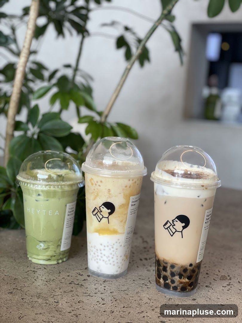 Three plastic cups of bubble tea are arranged on a textured surface. The cup on the left is green and has the logo "HEYTEA" printed on it. The middle cup is a light brown color with yellow and white layers, and appears to contain tapioca pearls. The cup on the right is a creamy brown color with a white foam topping and a layer of tapioca pearls at the bottom. A plant is visible in the background, along with a blurred interior space.