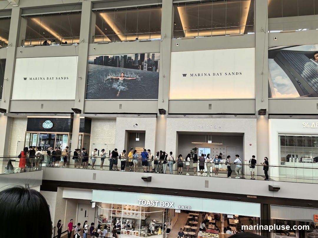 A view of the interior of a shopping mall with several storefronts visible. Large advertisements for Marina Bay Sands are displayed on the upper level. Below, there are shops including HEYTEA and TOAST BOX, with people queuing outside HEYTEA.