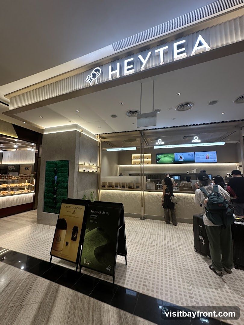 The exterior of a HEYTEA shop with a prominent illuminated sign. There are two A-frame signs in front of the shop, one advertising "BOBO TO THE WORLD" and the other "MATCHA ZEN + FOOD MENU". Several customers are visible inside and queuing.