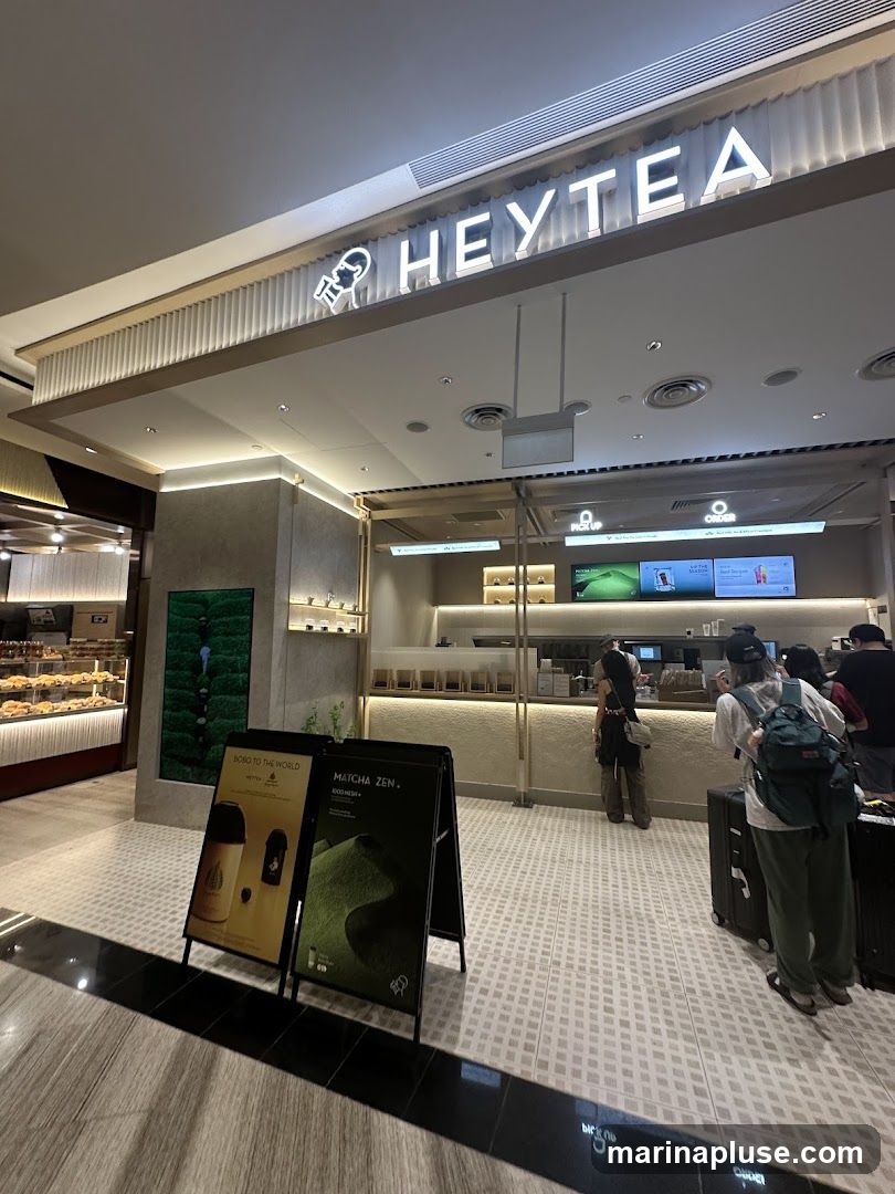 The exterior of a HEYTEA shop with a prominent illuminated sign. There are two A-frame signs in front of the shop, one advertising "BOBO TO THE WORLD" and the other "MATCHA ZEN + FOOD MENU". Several customers are visible inside and queuing.