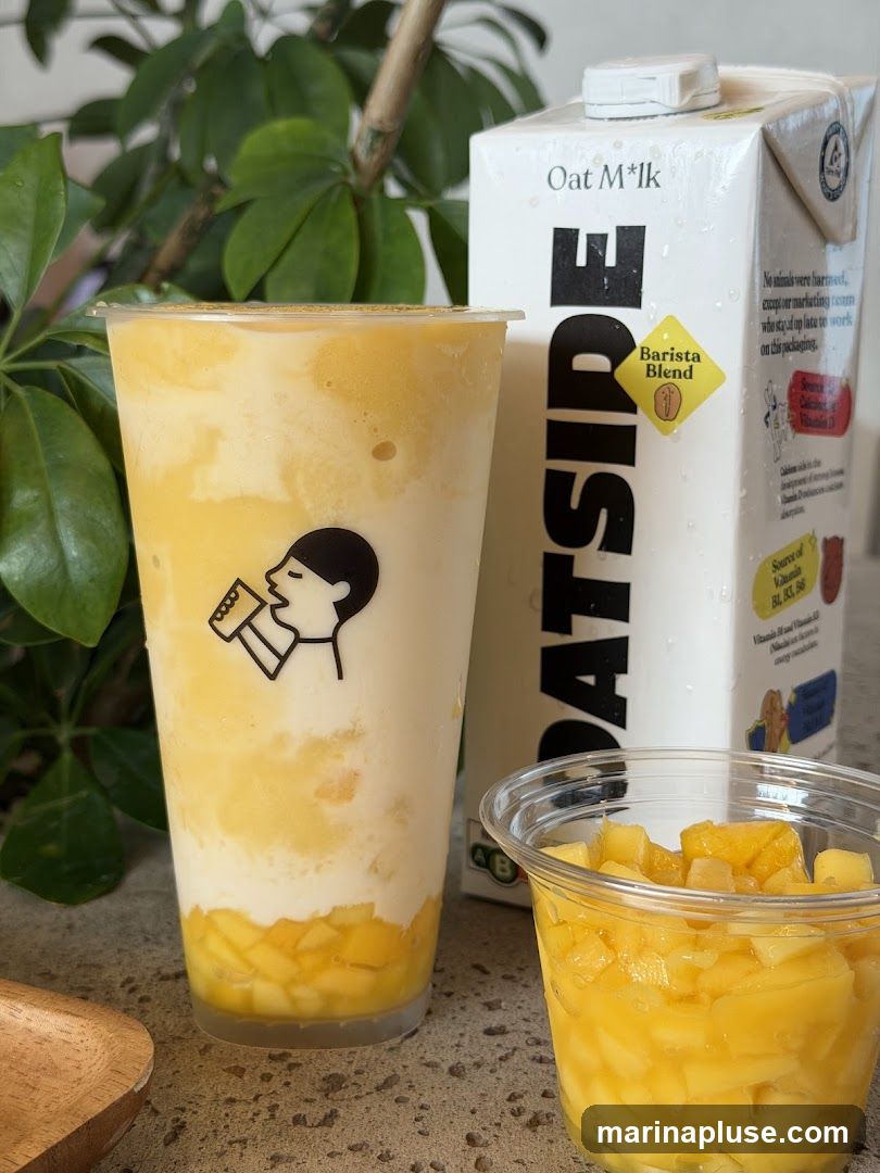A close-up shot of a layered mango drink in a clear plastic cup, with diced mangoes at the bottom and a creamy white layer above. A carton of Oat M*lk, labeled "Barista Blend," stands behind the drink. A small plastic cup filled with diced mangoes is also visible. The drink cup features a black and white logo of a person drinking.
