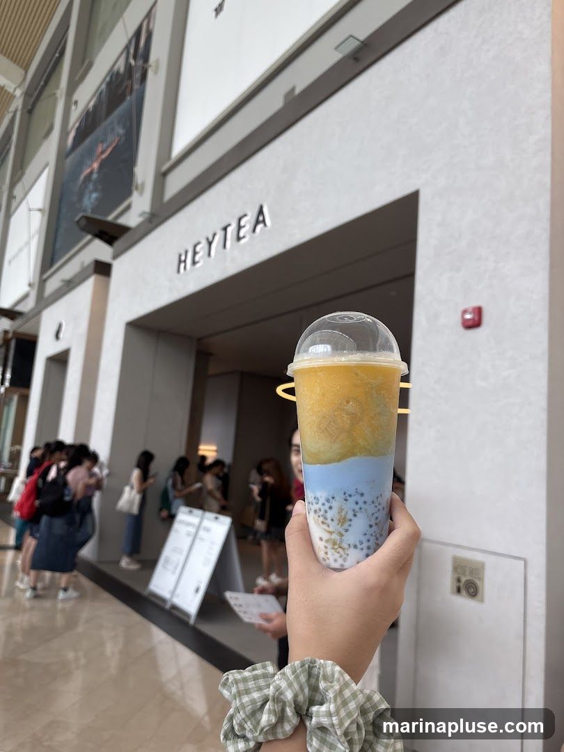 A person holds a layered beverage in front of a HEYTEA store. The store's name is visible above the entrance. In the background, people are queuing and there are large posters on the wall.