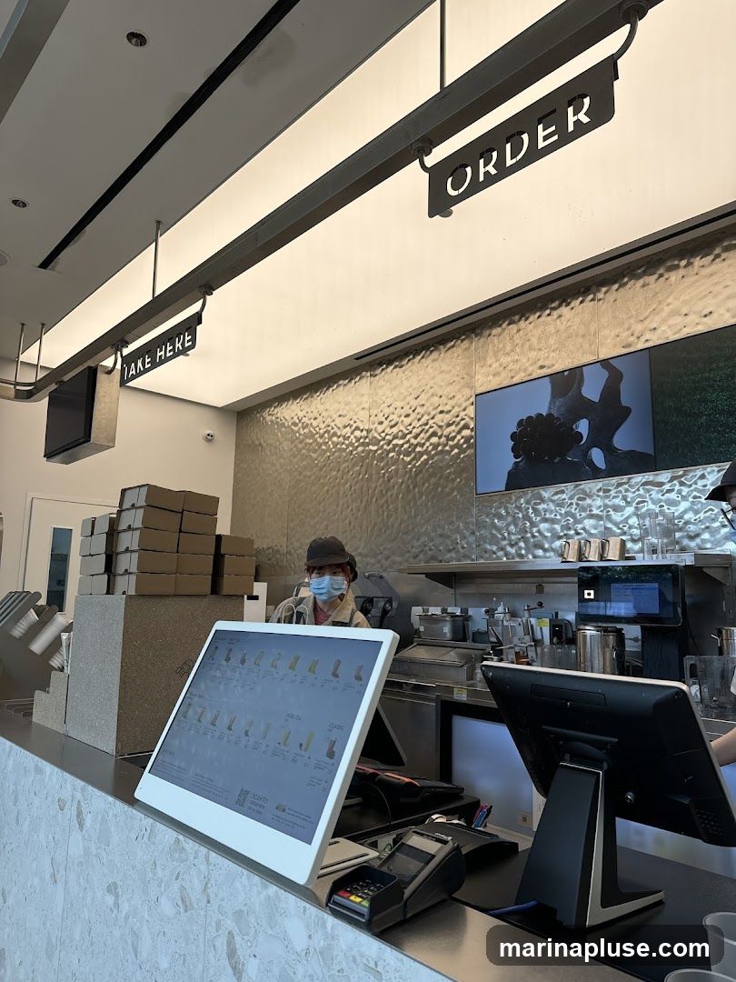A view of a counter with a digital menu displayed on a touchscreen. Signs for "ORDER" and "TAKE HERE" hang above. A person wearing a mask and hat stands behind the counter. Stacks of cardboard boxes are visible to the left.