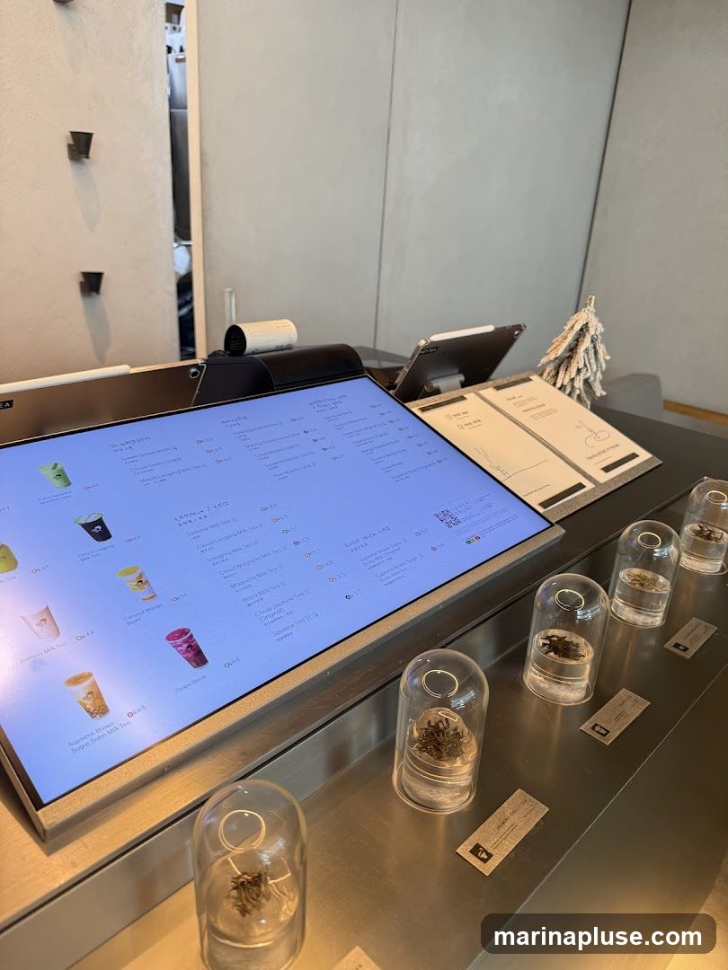 A digital menu displaying various tea options with prices. Small glass domes containing tea leaves are displayed in front of the menu.