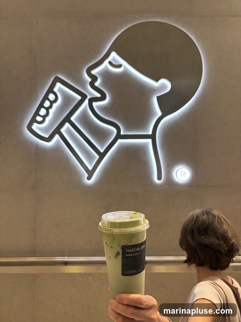 A person holds up a tall plastic cup filled with a green matcha drink. The cup has a clear lid and a black label that reads "MATCHA ZEN" and "1000 MESH". Behind the drink, a large, illuminated logo is visible on a grey wall. The logo is a stylized outline of a person's profile drinking from a cup. A small circular logo with an 'R' is also visible to the right of the main logo.