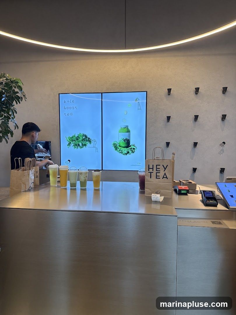 A modern beverage counter with a metallic finish displays several colorful drinks. Behind the counter, a large digital screen shows an advertisement for "kale boost tea" with images of kale and a jar. A person is visible behind the counter, appearing to prepare drinks. Several branded paper bags with the "HEY TEA" logo are placed on the counter. The overall ambiance is clean and contemporary, with minimalist decor and a prominent LED light fixture overhead.