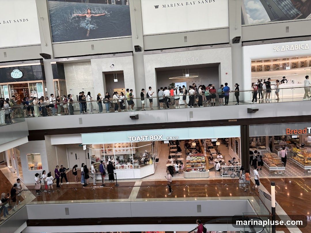 A multi-level shopping mall interior with several shops visible. On the upper level, there are signs for MARINA BAY SANDS, HEYTEA, and % ARABICA, with many people queuing. On the lower level, TOAST BOX and BreadTalk are visible, also with customers. The mall features modern architecture and a large open atrium.