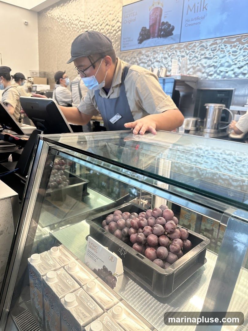 A masked staff member wearing an apron is seen behind a counter, interacting with a computer screen. In the foreground, a display case shows cartons of drinks and a crate of Kyoho grapes. A promotional poster for HEYTEA Original is visible in the background.