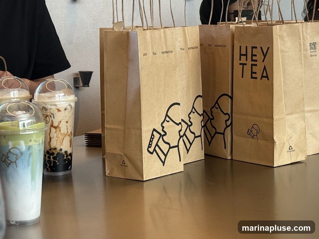 Several takeaway drinks with plastic lids are lined up on a counter. Behind them are several brown paper bags with handles. The bags have black line drawings of people drinking and the words "HEY TEA" printed on them.