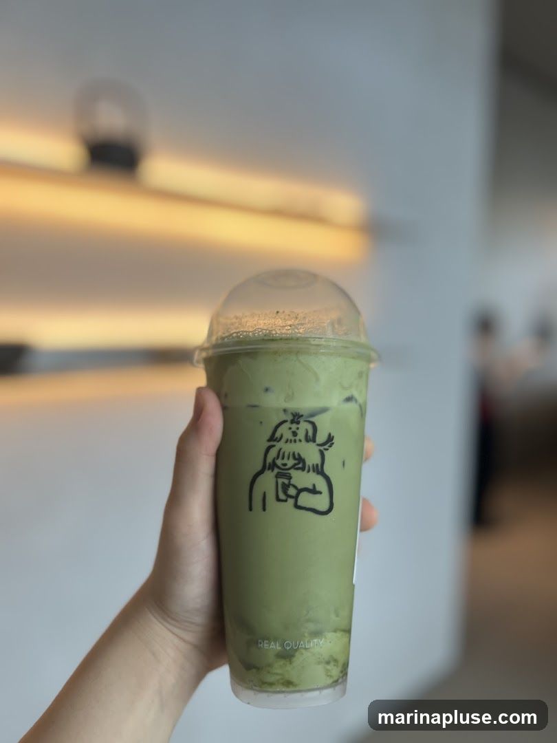 A hand holds a clear plastic cup filled with a green matcha drink. The cup has a cartoon illustration of a person with a dog on their head, holding a cup. Text at the bottom of the cup reads "REAL QUALITY". The background is blurred, showing warm lighting and a hint of an interior space.