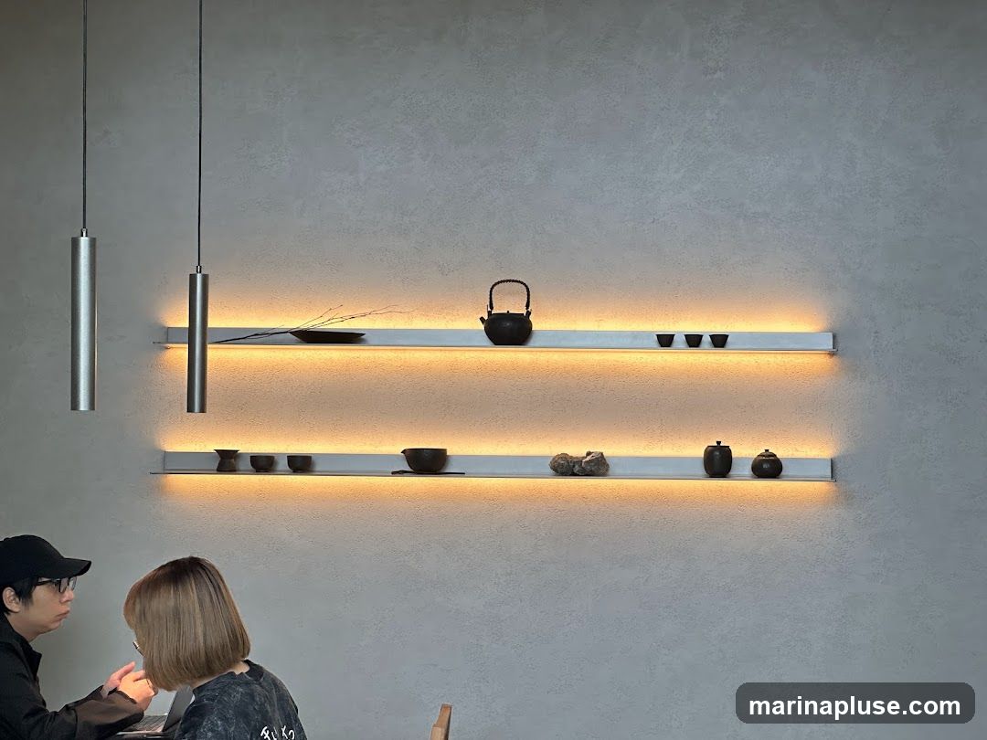 Two people are seated at a table in a minimalist interior. Above them, two long shelves are mounted on a textured gray wall, each illuminated by warm LED lighting. The top shelf displays a teapot, a small branch, and three cups. The bottom shelf holds decorative stones and two small lidded jars. Two cylindrical pendant lights hang from the ceiling on the left side of the image.