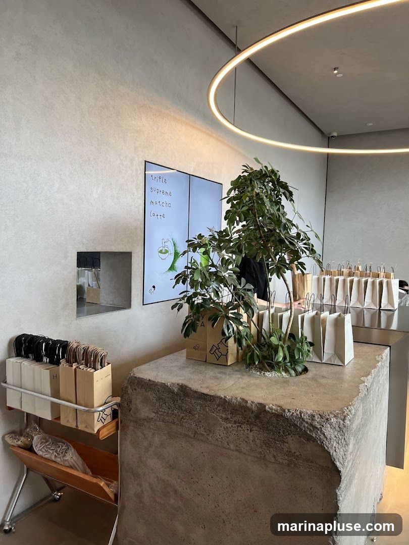 A modern cafe interior featuring a large digital screen displaying a menu with "Triple Supreme Matcha Latte" as a prominent item. The screen shows a graphic of matcha being prepared. In the foreground, a rough-hewn concrete counter is adorned with potted plants and several paper bags. To the left, a metal rack holds more paper bags and other items. The overall ambiance is minimalist and contemporary, with a circular light fixture overhead.