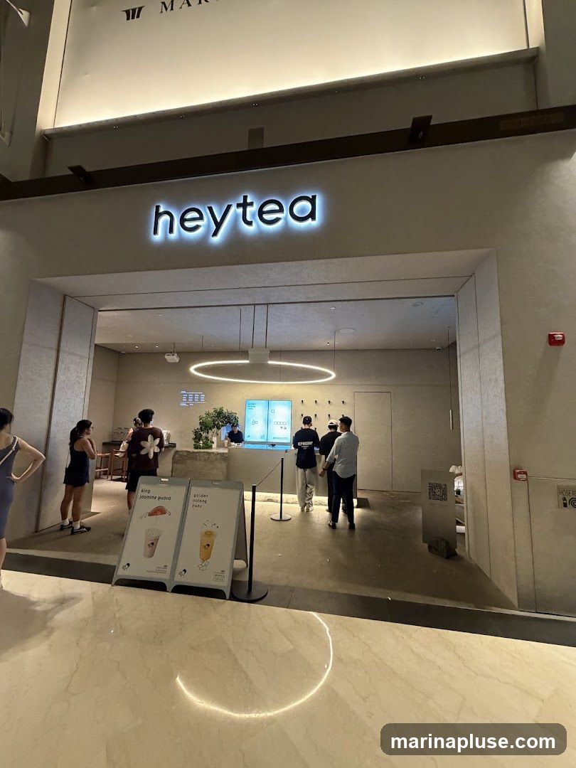The exterior of a modern bubble tea shop with the illuminated "heytea" logo above the entrance. Inside, customers are seen ordering at the counter. Two A-frame signs are placed outside the entrance, displaying images of drinks and their names.