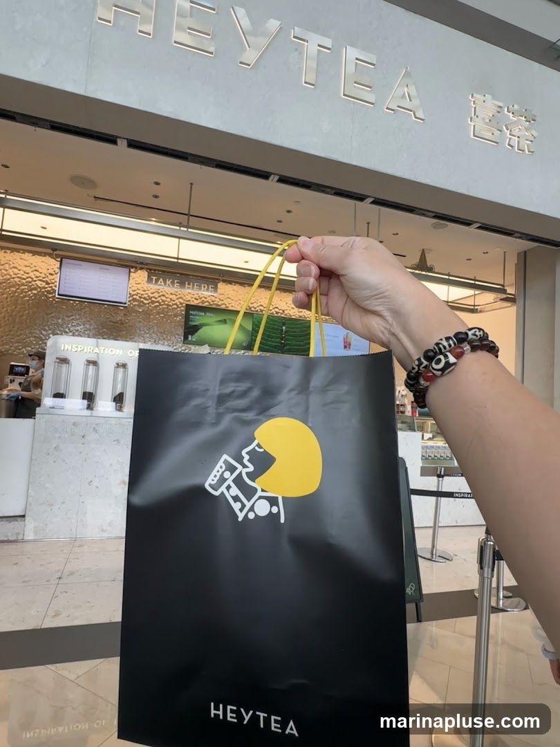 A person holds a black shopping bag with the Heytea logo in front of a Heytea store. The store has a modern facade with the Heytea logo prominently displayed in silver lettering above the entrance. Inside, a counter with a digital menu board is visible, along with queuing stanchions.