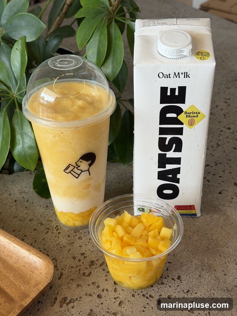 A tall plastic cup filled with a layered mango smoothie, topped with a clear dome lid. Next to it, a small clear plastic cup contains diced mango. A carton of Oat M*lk Barista Blend stands behind the drinks. A wooden tray is partially visible on the left.