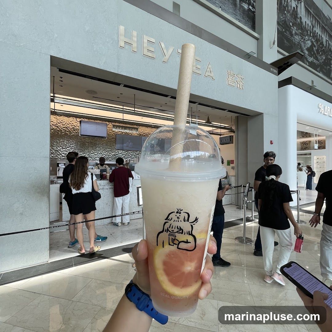 A person holds a beverage in front of a Hey Tea store. The store has a modern, clean design with the brand name "HEY TEA" displayed prominently in gold lettering above the entrance. Several customers are visible inside and in line.