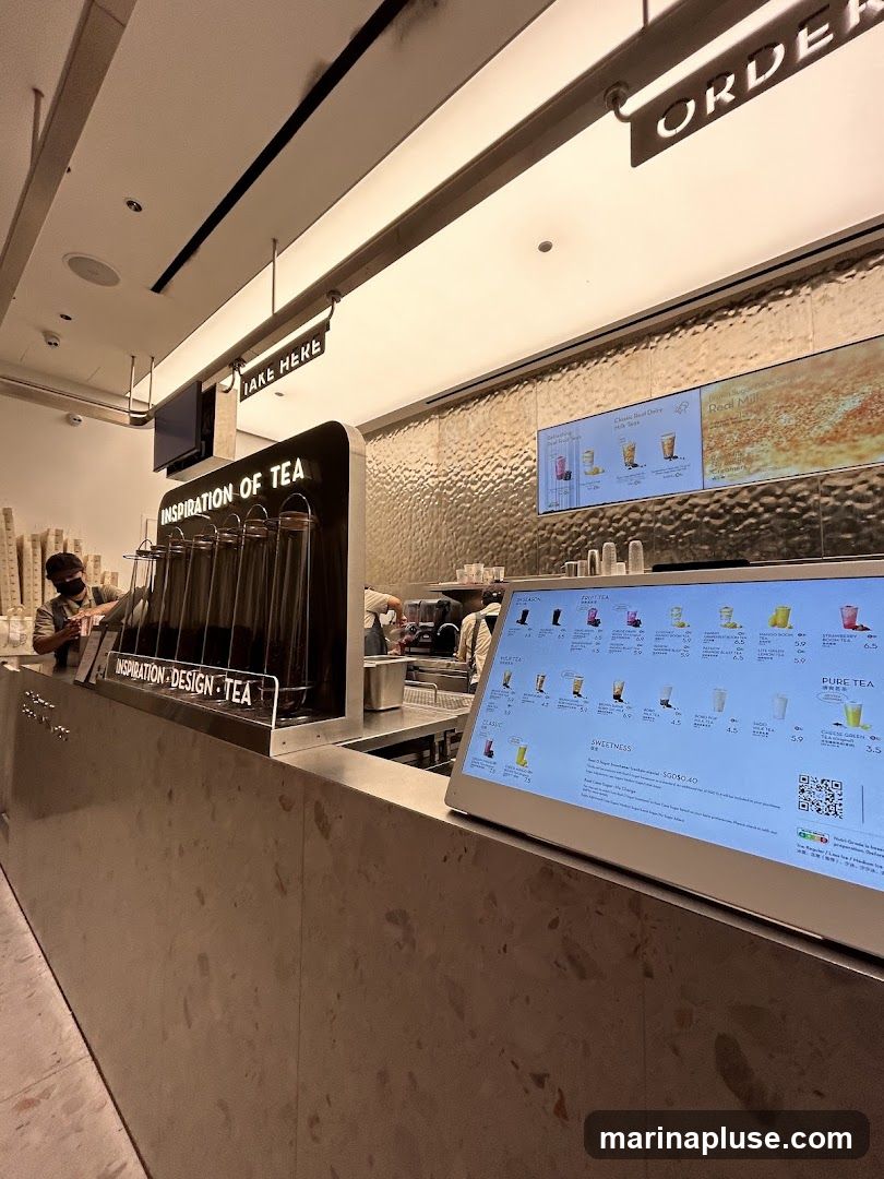 A view of a tea shop counter with digital menus displaying various drinks and options. Signs for "ORDER" and "TAKE HERE" are visible. The counter has a textured surface, and behind it, staff members are preparing drinks. The digital menu shows categories like "FRUIT TEA", "CLASSIC", and "PURE TEA" with prices.