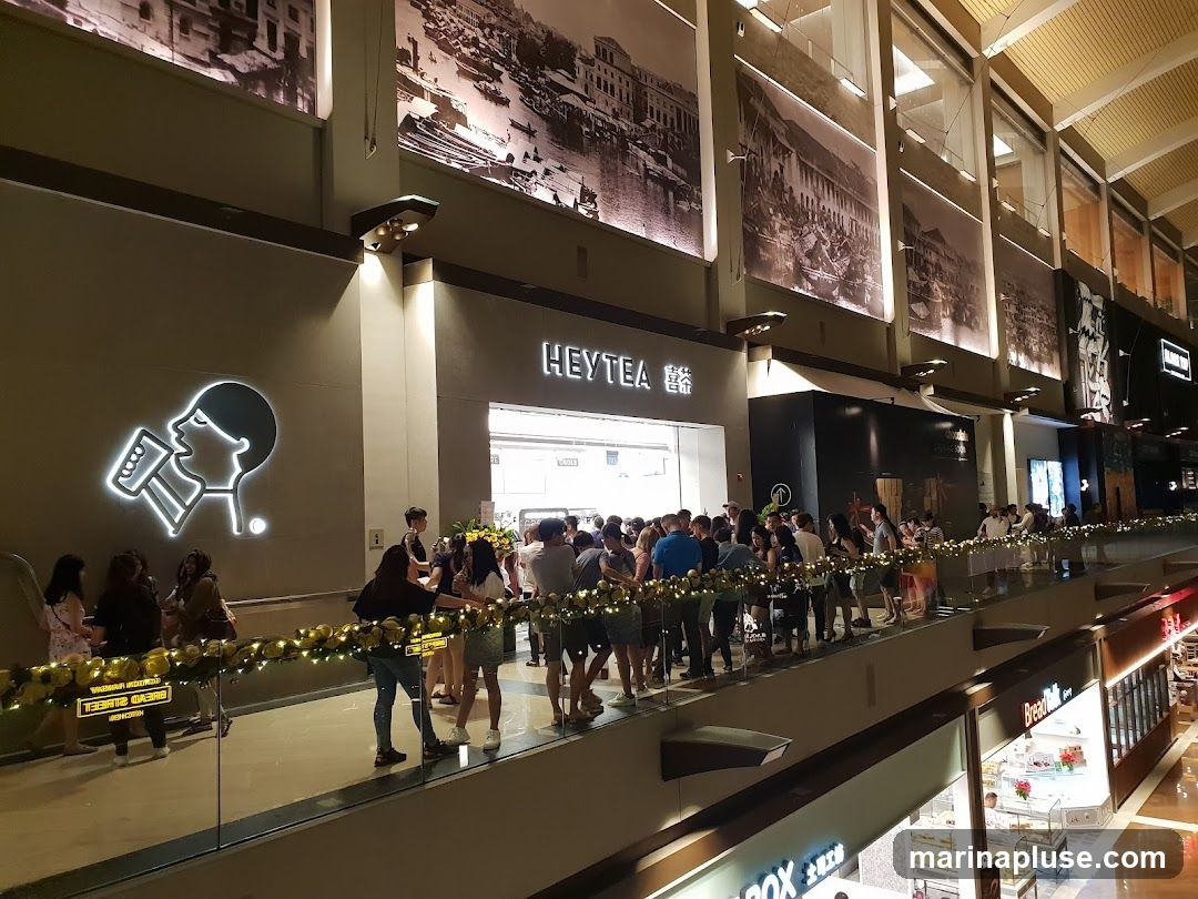 A busy HEYTEA shop with a long queue of customers. The shop has a prominent neon sign of a person drinking from a cup. Large historical-themed murals are displayed on the wall above the shop.