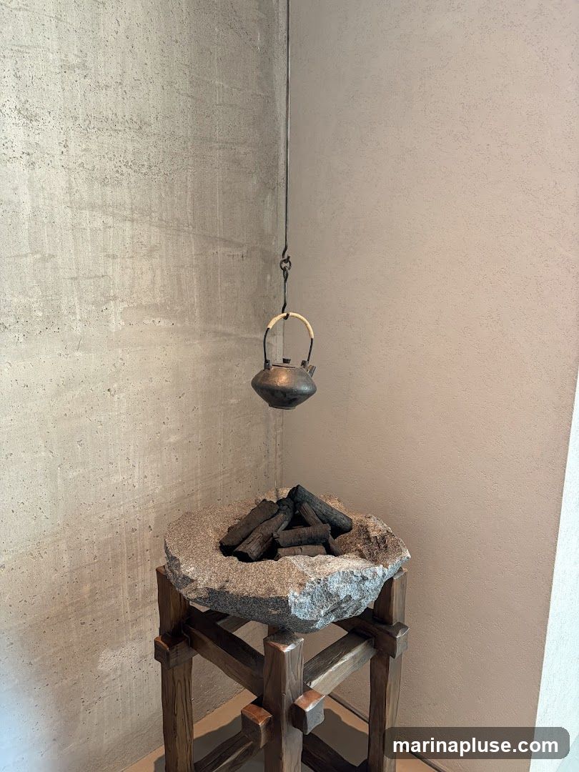A decorative display featuring a small, dark metal teapot suspended by a wire above a stone basin filled with charcoal pieces. The stone basin is supported by a wooden stand with a rustic design. The background consists of textured concrete walls.
