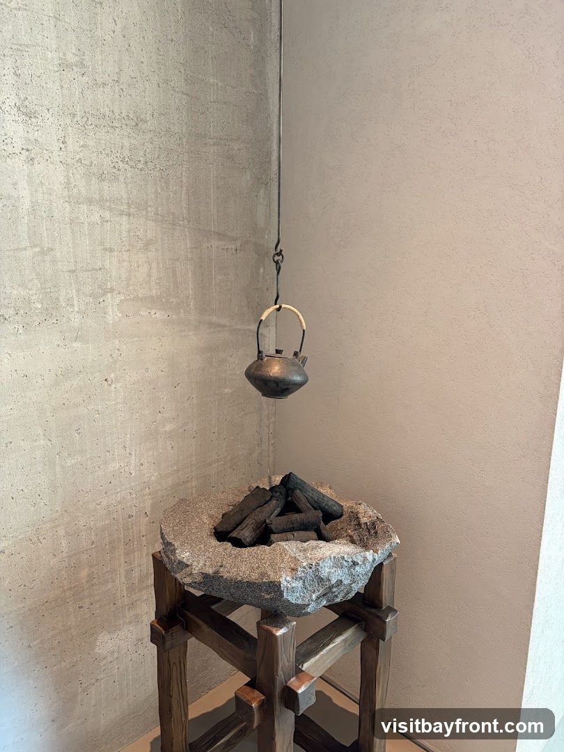 A decorative display featuring a small, dark metal teapot suspended by a wire above a stone basin filled with charcoal pieces. The stone basin is supported by a wooden stand with a rustic design. The background consists of textured concrete walls.