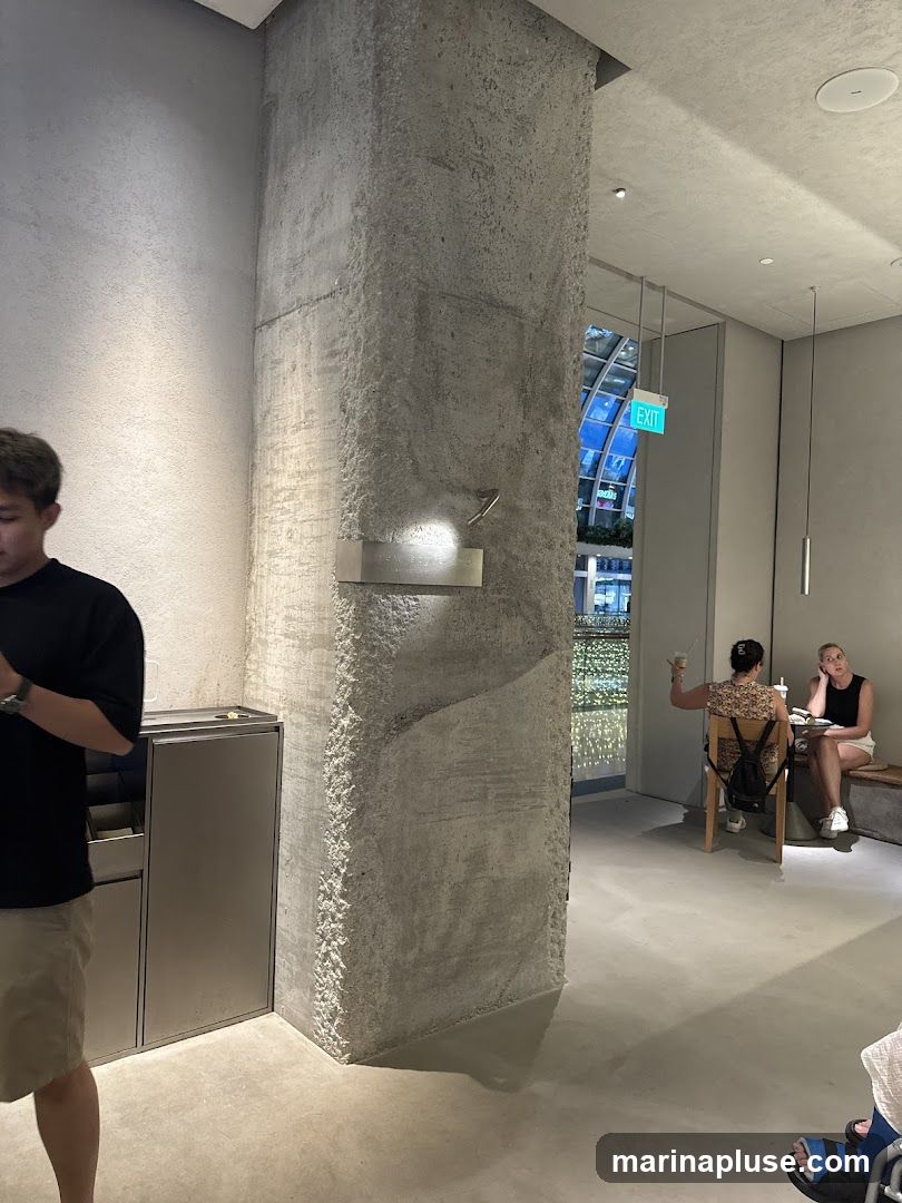 The image shows the interior of a cafe with a rough concrete pillar in the foreground. A man in a black t-shirt and beige shorts stands to the left. In the background, two women are seated at tables. One woman is gesturing while holding a drink, and the other is reading a book. An exit sign is visible in the distance. The overall ambiance is minimalist and contemporary.