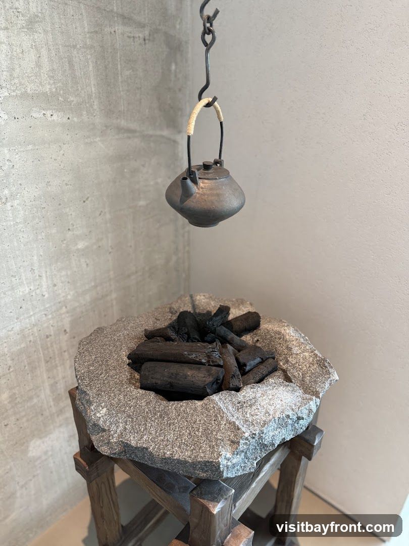 A rustic display featuring a dark, textured kettle suspended by a metal chain above a rough, stone basin filled with charcoal pieces. The basin rests on a wooden stand. The background consists of textured concrete and plain walls.