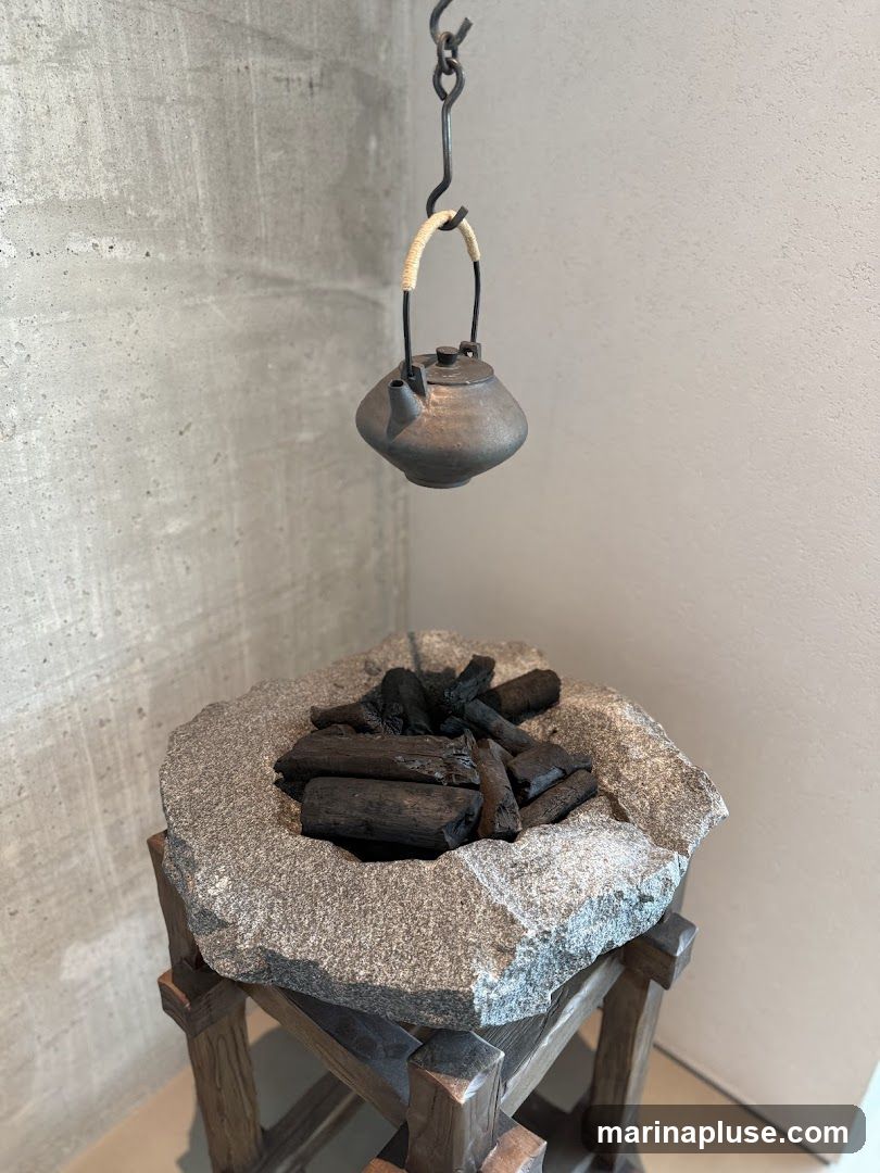 A rustic display featuring a dark, textured kettle suspended by a metal chain above a rough, stone basin filled with charcoal pieces. The basin rests on a wooden stand. The background consists of textured concrete and plain walls.