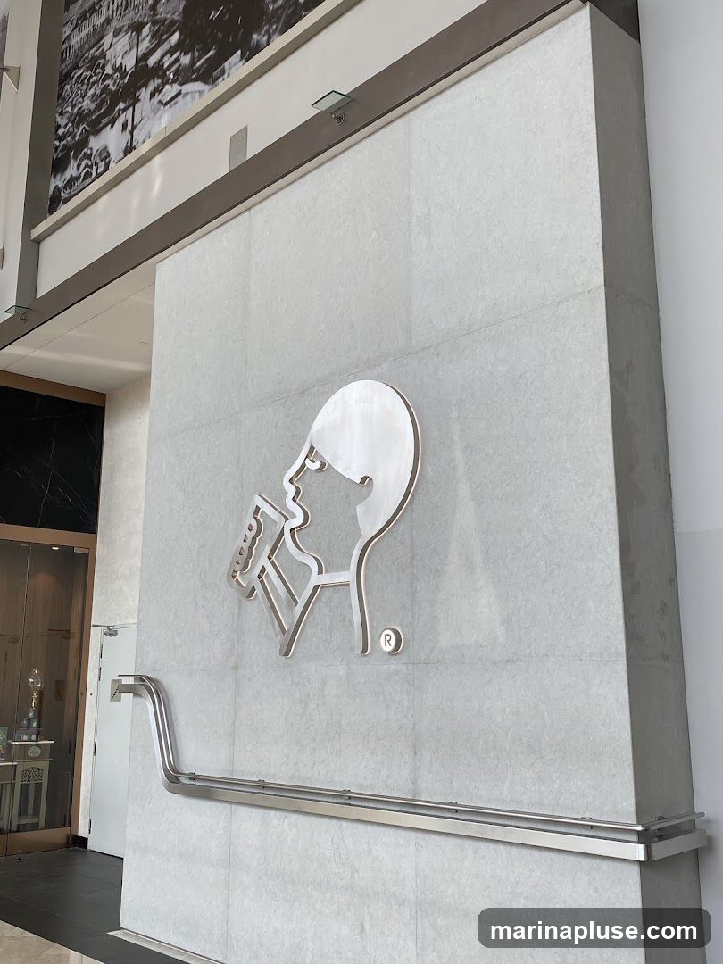 A large, metallic logo of a person holding a phone is mounted on a light gray tiled wall. Above the logo, a large black and white mural depicts a cityscape. A silver handrail runs horizontally across the lower portion of the wall. To the left, a glass door leads into an interior space with a decorative display.
