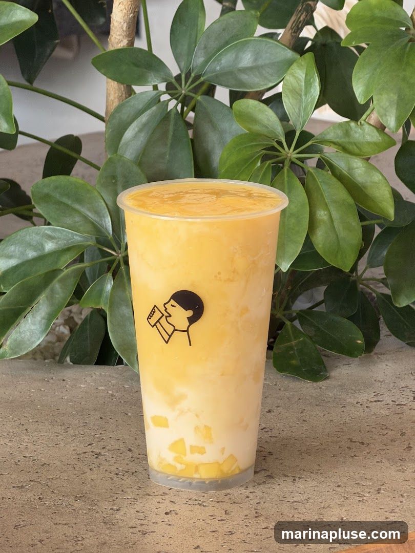 A close-up shot of a mango drink in a clear plastic cup with a black logo of a person drinking. The drink appears to have layers of mango puree, milk, and diced mango pieces at the bottom. The cup is placed on a textured surface, with lush green foliage in the background.