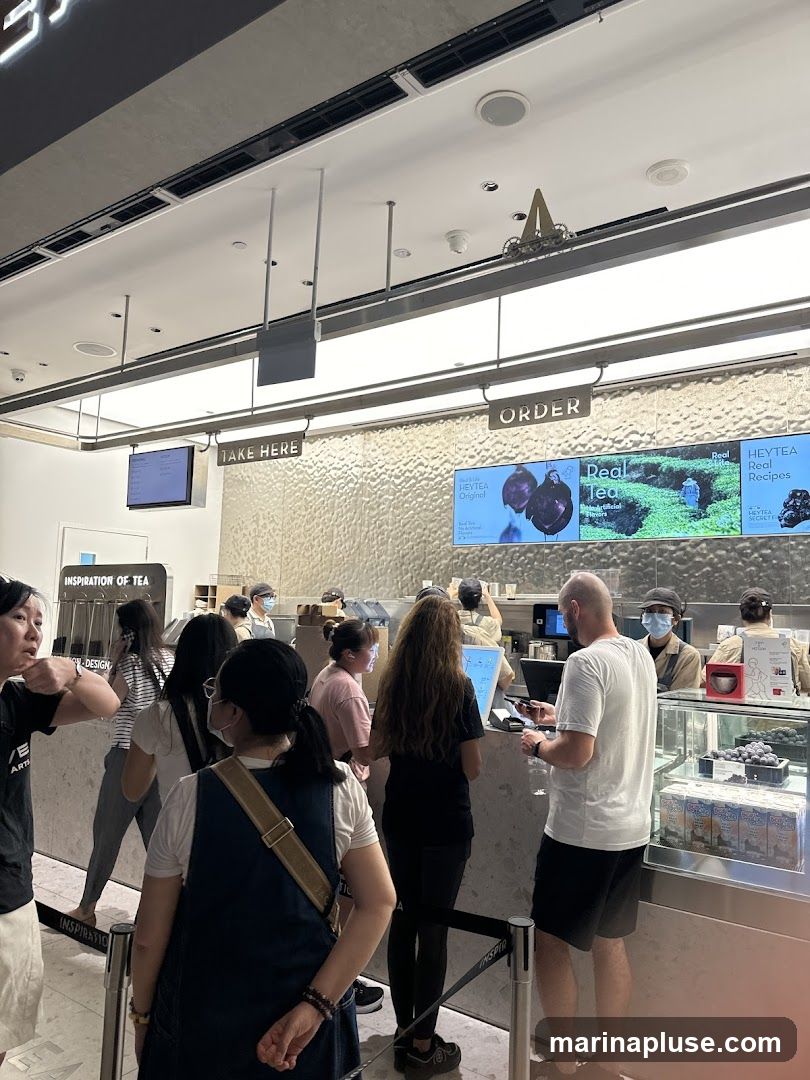 A busy tea shop counter with customers queuing to order and take their drinks. Digital screens display menu items and promotions. Staff members are visible behind the counter preparing orders.