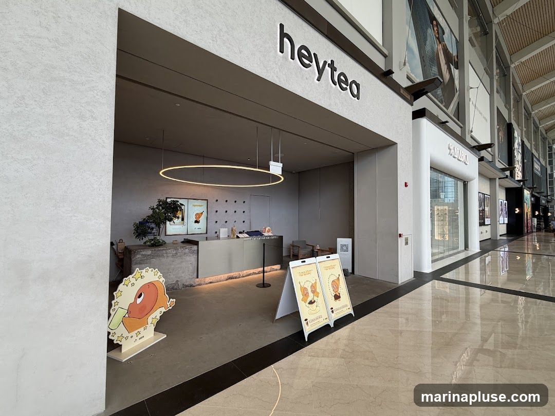 The exterior of a Heytea store with its prominent signage above the entrance. The store features a modern design with a counter visible inside. Two A-frame signs with cartoon characters are placed outside the entrance. To the right, the entrance to % Arabica is visible.