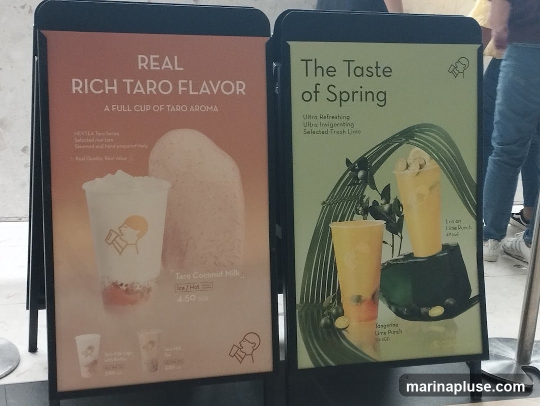Two A-frame signs display advertisements for beverages. The left sign promotes "REAL RICH TARO FLAVOR" with a description of HEYTEA Taro Series, featuring Taro Coconut Milk (Ice/Hot) for 4.50 SGD, Taro Milk Sage with Banday for 3.80, and Taro Milk for 3.80. The right sign advertises "The Taste of Spring" with "Ultra Refreshing Ultra Invigorating Selected Fresh Lime" drinks, including Tangerine Lime Punch for 5.4 SGD and Lemon Lime Punch for 4.9 SGD.