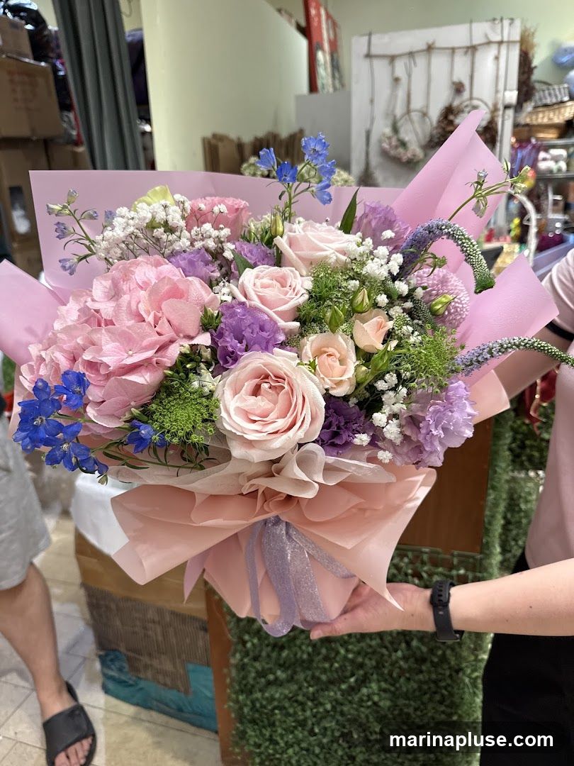 A person is holding a large bouquet of flowers. The bouquet contains pink hydrangeas, pink roses, purple lisianthus, and blue delphiniums, with accents of baby's breath and other greenery. The flowers are wrapped in pink and peach paper and tied with a purple ribbon. In the background, there are shelves with various items, including boxes, baskets, and decorative elements.